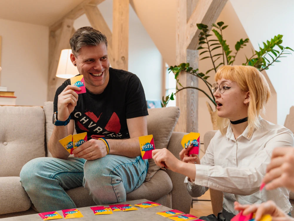 Cilvēki spēlē dinamisko kāršu spēli Piles! ar krāsainām kārtīm, mēģinot savākt apģērbu komplektus uz galda.
