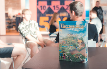 Carcassonne galda spēles kaste uz galda, fonā redzami cilvēki spēļu pasākumā.