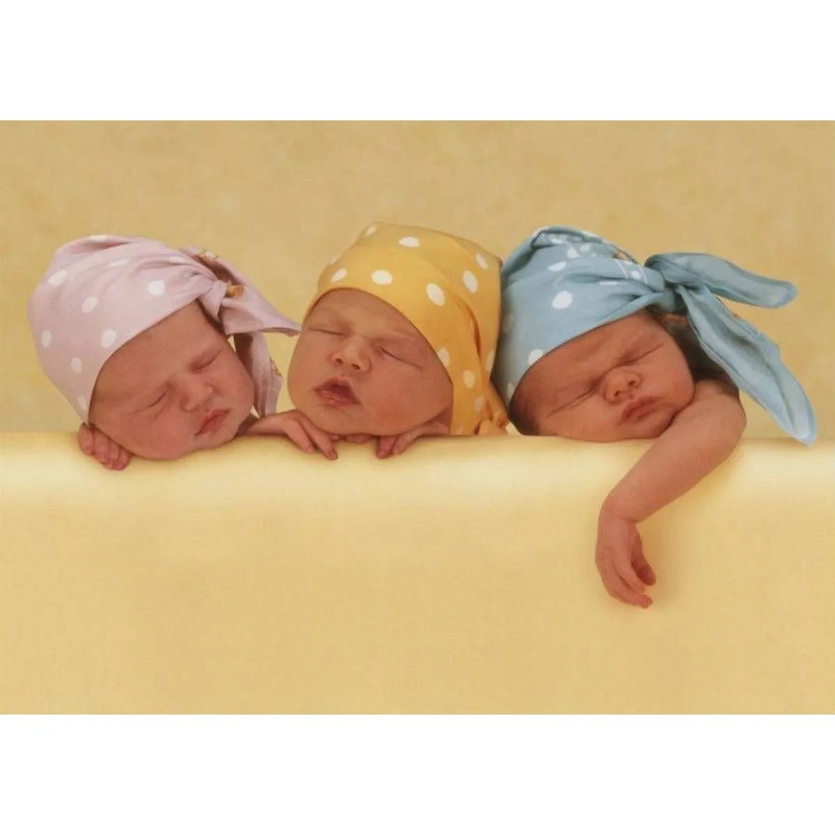 Anne Geddes 1000 gabaliņu puzle ar trim mazuļiem krāsainās cepurītēs uz dzeltena fona.