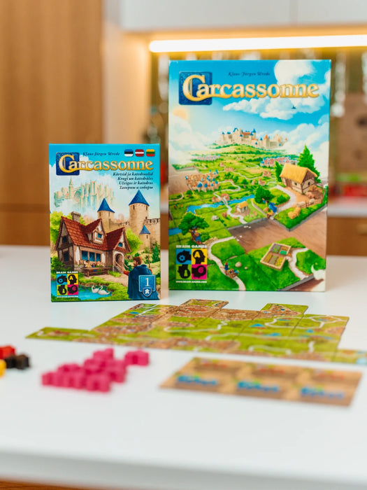 Carcassonne Krogi un katedrāles paplašinājuma kaste ar spēles lauciņiem un rozā koka figūriņām uz galda.