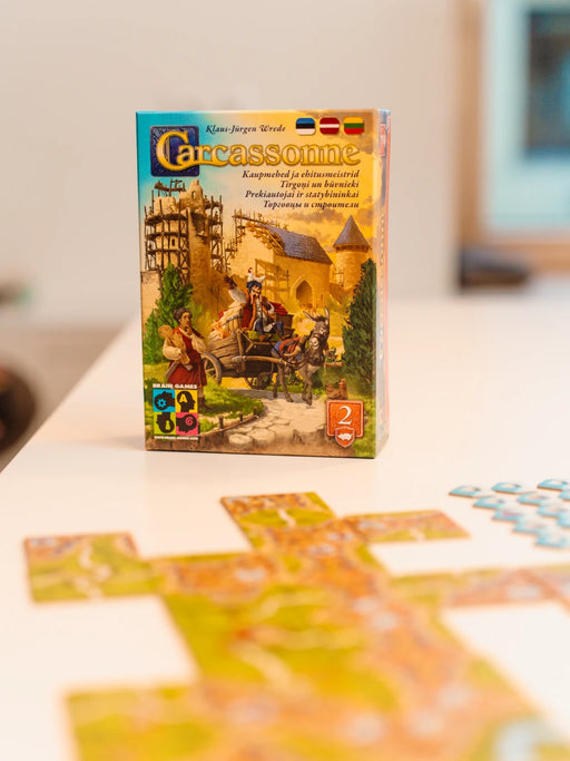 Carcassonne paplašinājuma Tirgoņi un būvnieki kastes vāks ar ilustrāciju un izkārtoti spēles lauciņi uz galda.
