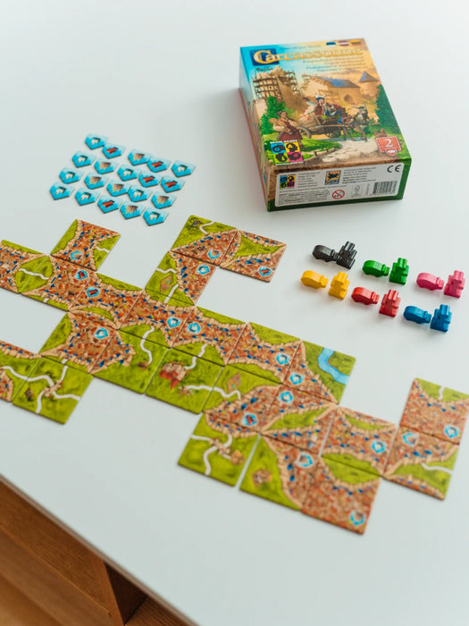 Carcassonne paplašinājuma kaste, krāsaini koka būvnieki, sivēni un izkārtoti spēles lauciņi uz balta galda.