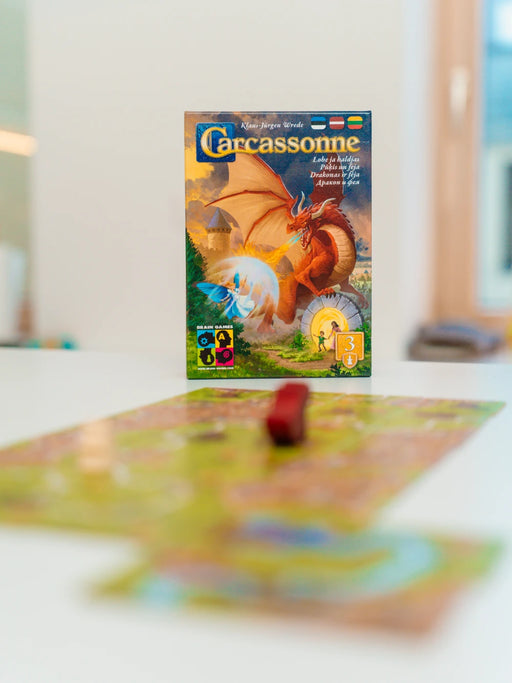 Carcassonne paplašinājuma Pūķis un feja kastes vāks ar sarkanu pūķi un feju uz galda ar spēles laukumu.