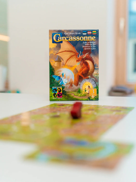 Carcassonne paplašinājuma Pūķis un feja kastes vāks ar sarkanu pūķi un feju uz galda ar spēles laukumu.