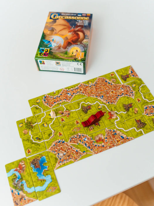 Carcassonne paplašinājuma Pūķis un feja kaste ar izliktiem kartona laukuma elementiem, sarkanu pūķa figūriņu un spēles kauliņiem.