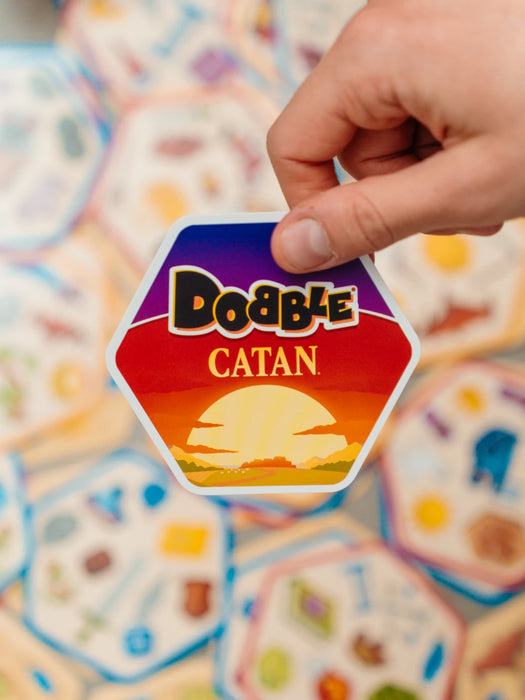 Roka tur sešstūrainu Dobble CATAN spēles kārti ar saulrieta dizainu uz citu kāršu fona.