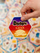 Roka tur sešstūrainu Dobble CATAN spēles kārti ar saulrieta dizainu uz citu kāršu fona.