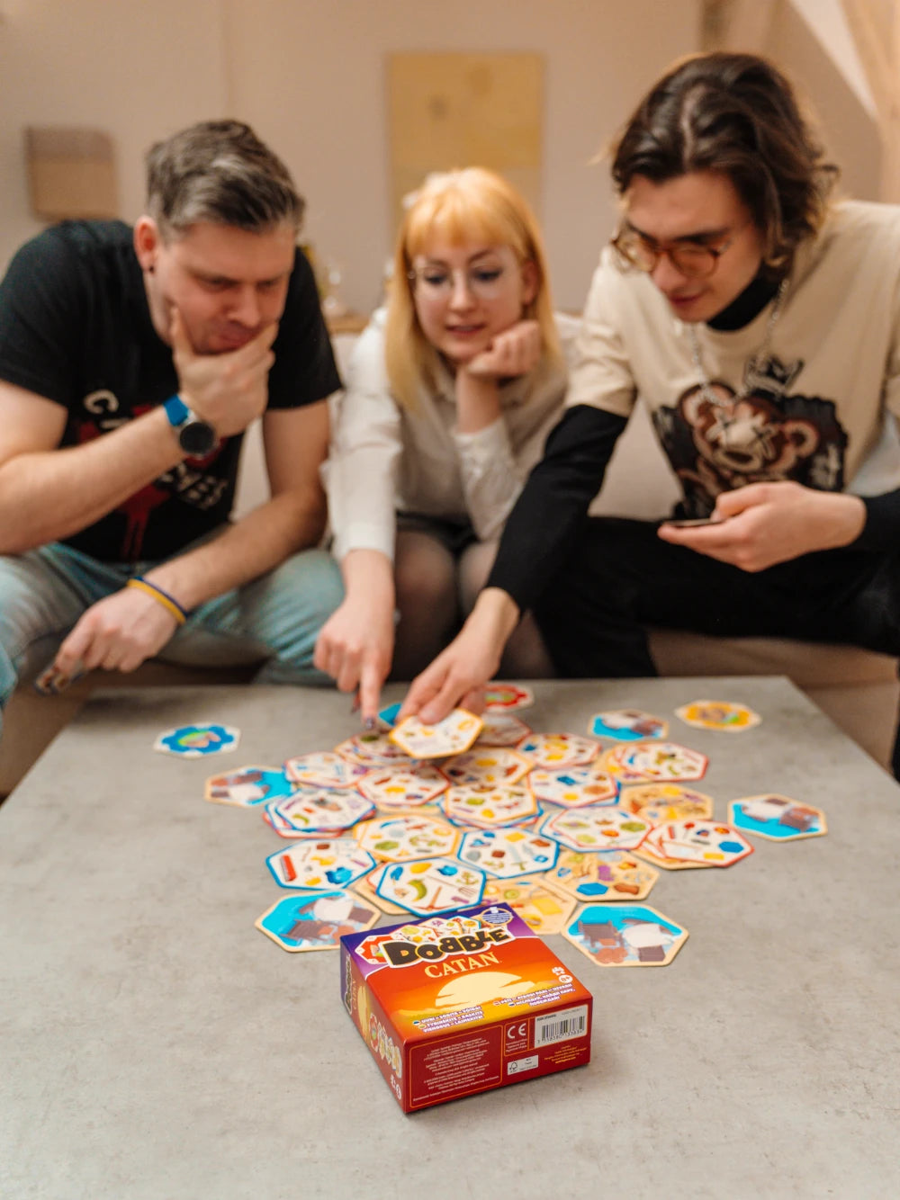 Trīs cilvēki spēlē Dobble CATAN galda spēli ar sešstūrainām kārtīm un krāsainiem simboliem uz galda.