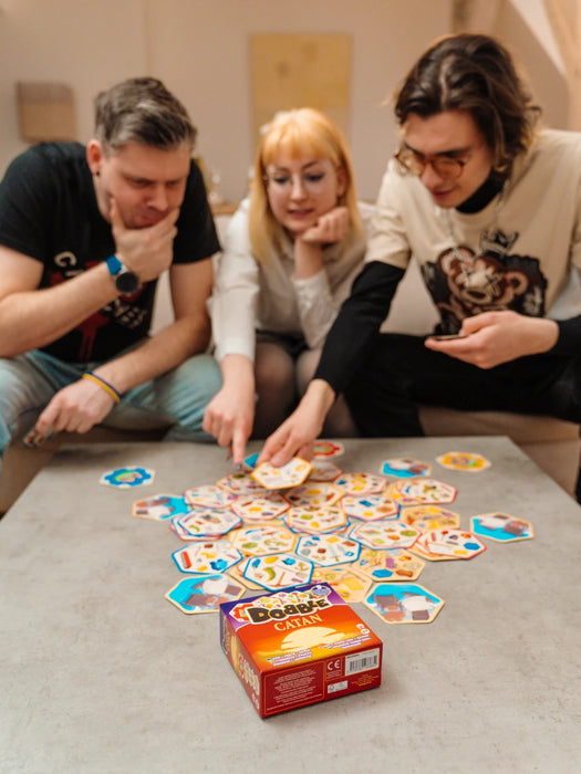 Trīs cilvēki spēlē Dobble CATAN galda spēli ar sešstūrainām kārtīm un krāsainiem simboliem uz galda.
