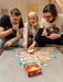 Trīs cilvēki spēlē Dobble CATAN galda spēli ar sešstūrainām kārtīm un krāsainiem simboliem uz galda.