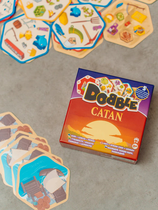 Dobble CATAN galda spēles kaste ar sešstūrainām kārtīm, kurās attēloti dažādi spēles simboli uz pelēka fona.