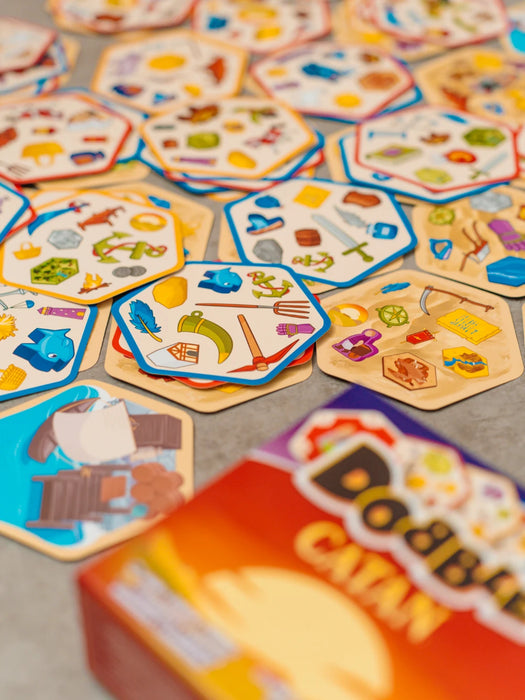 Dobble CATAN galda spēle ar sešstūrainām kārtīm, uz kurām attēloti krāsaini Katanas salas simboli un priekšmeti.