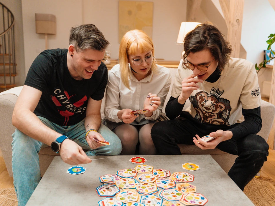 Trīs draugi spēlē Dobble CATAN galda spēli ar krāsainām sešstūrainām kārtīm uz galda mājīgā istabā.