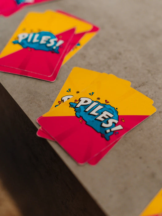 Spēles Piles! kārtis ar košu dzeltenu un rozā dizainu un zilu logotipu uz pelēkas virsmas.