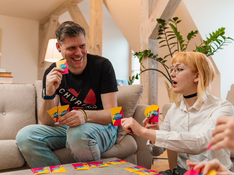 Cilvēki spēlē dinamisko kāršu spēli Piles! ar krāsainām kārtīm, mēģinot savākt apģērbu komplektus uz galda.