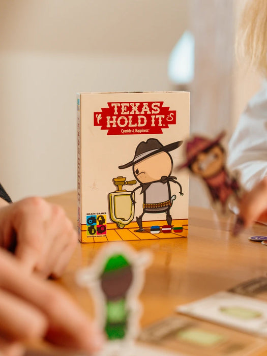 Texas Hold it! kāršu spēles iepakojums ar kovboju un pisuāru uz galda, blakus izkārtotas spēles kārtis un figūriņas.