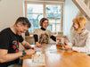 Trīs draugi smejoties spēlē Texas Hold It kāršu spēli pie koka galda ar spēles kastīti un kārtīm.