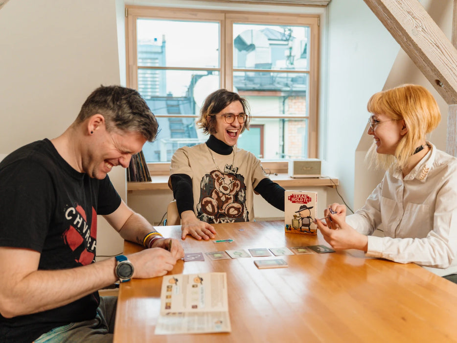 Trīs draugi smejoties spēlē Texas Hold It kāršu spēli pie koka galda ar spēles kastīti un kārtīm.