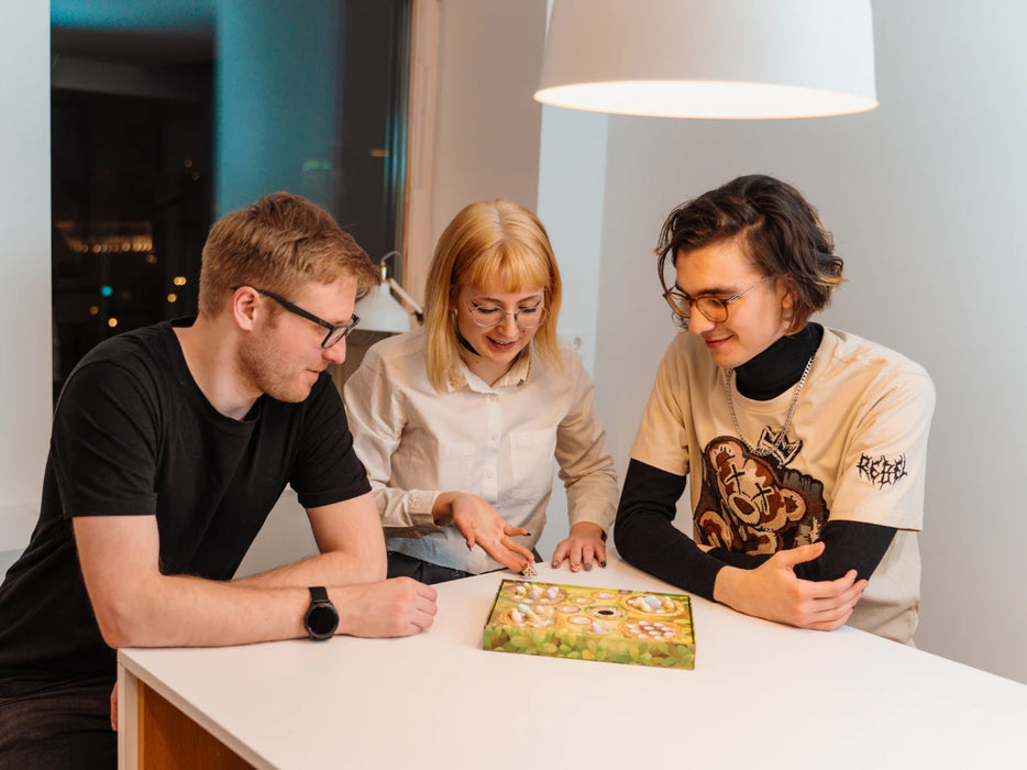 Trīs jaunieši spēlē galda spēli Tweet Tweet ar krāsainām olām un kauliņiem uz balta galda.